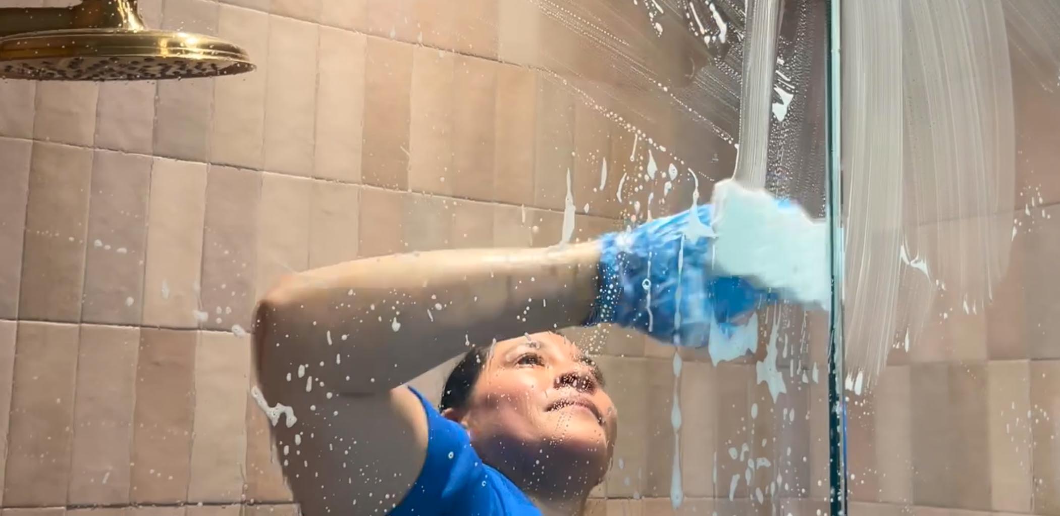 A person wearing blue gloves performs deep cleaning in Georgia on a glass shower door with a sponge, surrounded by soap suds and tiled walls.