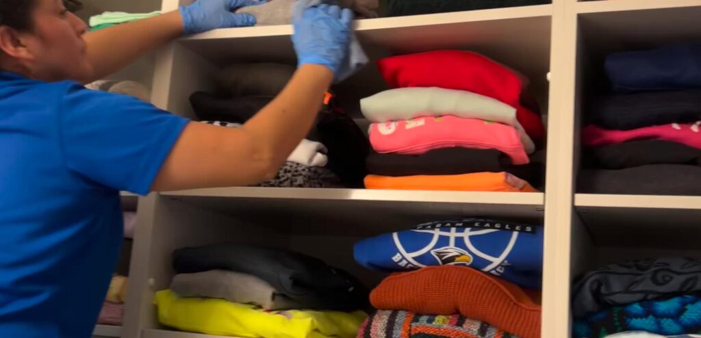 A person wearing blue gloves is cleaning shelves above stacks of folded clothes in a closet.