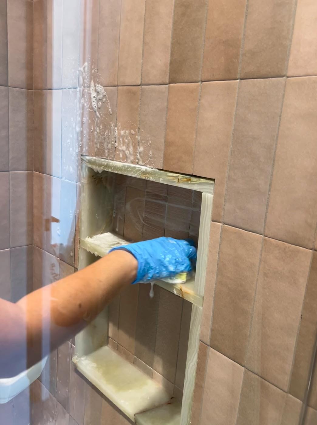 A person wearing a blue glove scrubs a built-in shower shelf with cleaning foam on tan tiled walls.