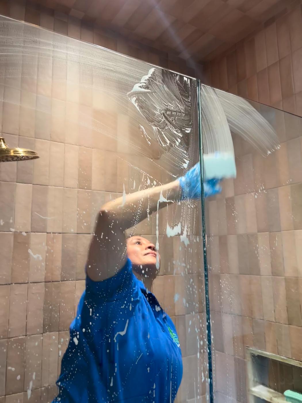 A person wearing blue gloves and a blue shirt is cleaning a glass shower door with a sponge inside a tiled bathroom. To learn more, contact CK Cleaning.