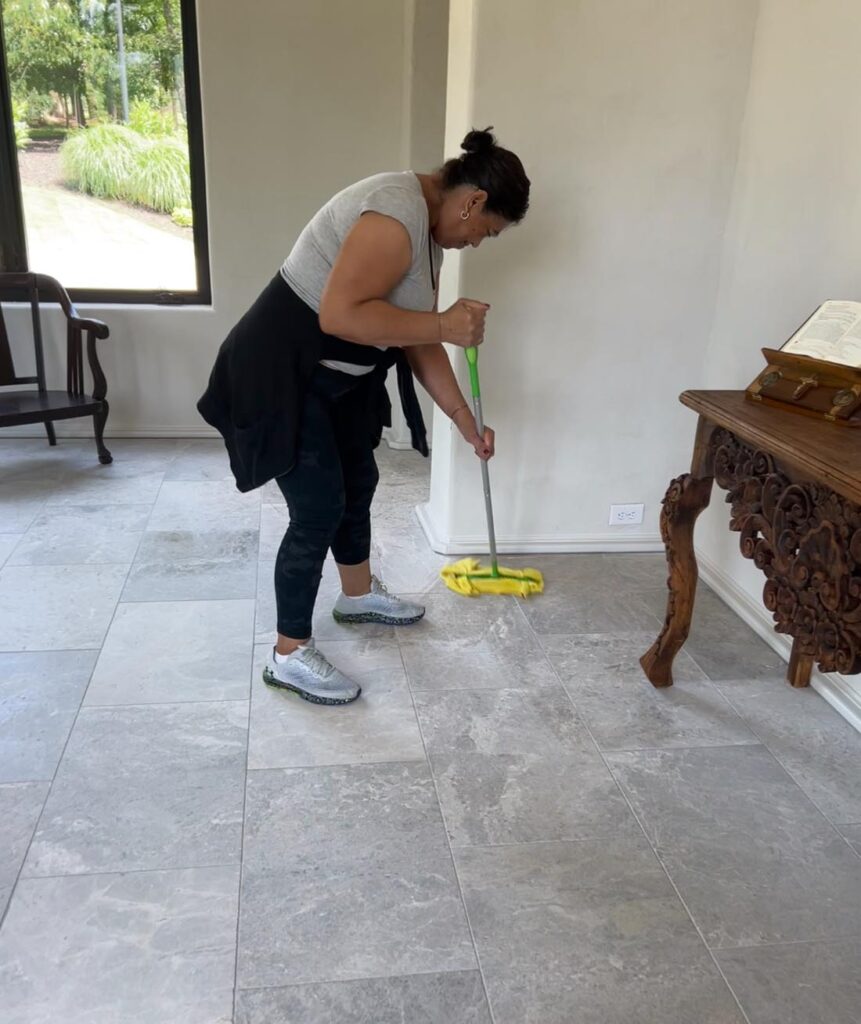 A woman is mopping a tiled floor in a room with a window, a wooden chair, and a carved wooden table.