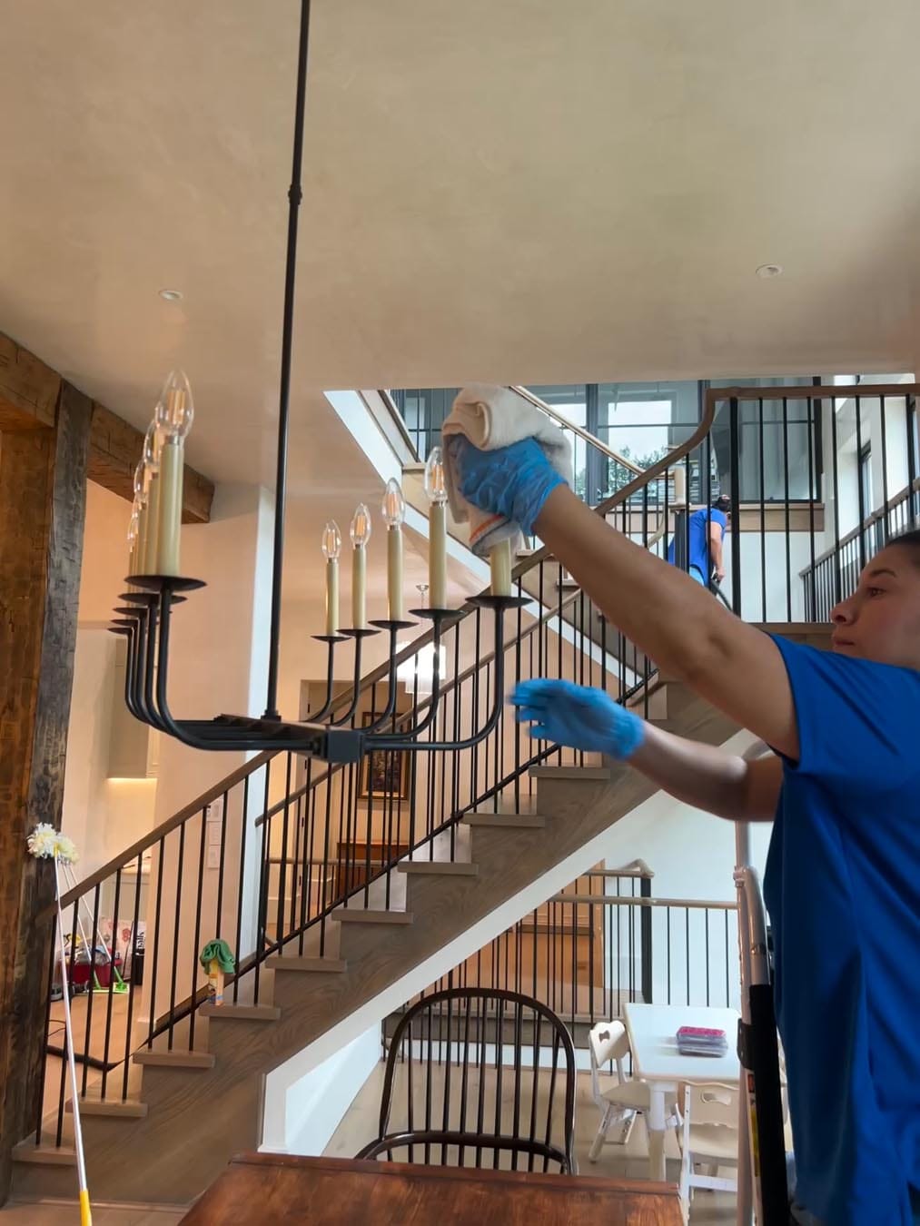 A person wearing blue gloves cleans a black chandelier in a modern home with wooden stairs and a minimalist interior.