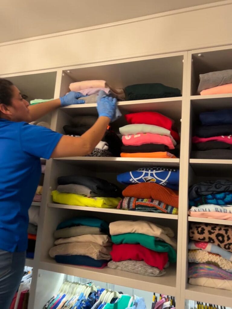 Cleaning services in Georgia: A CK Cleaning staff wearing blue gloves and a blue shirt is organizing folded clothes on the top shelf of a closet with multiple stacks of clothing.