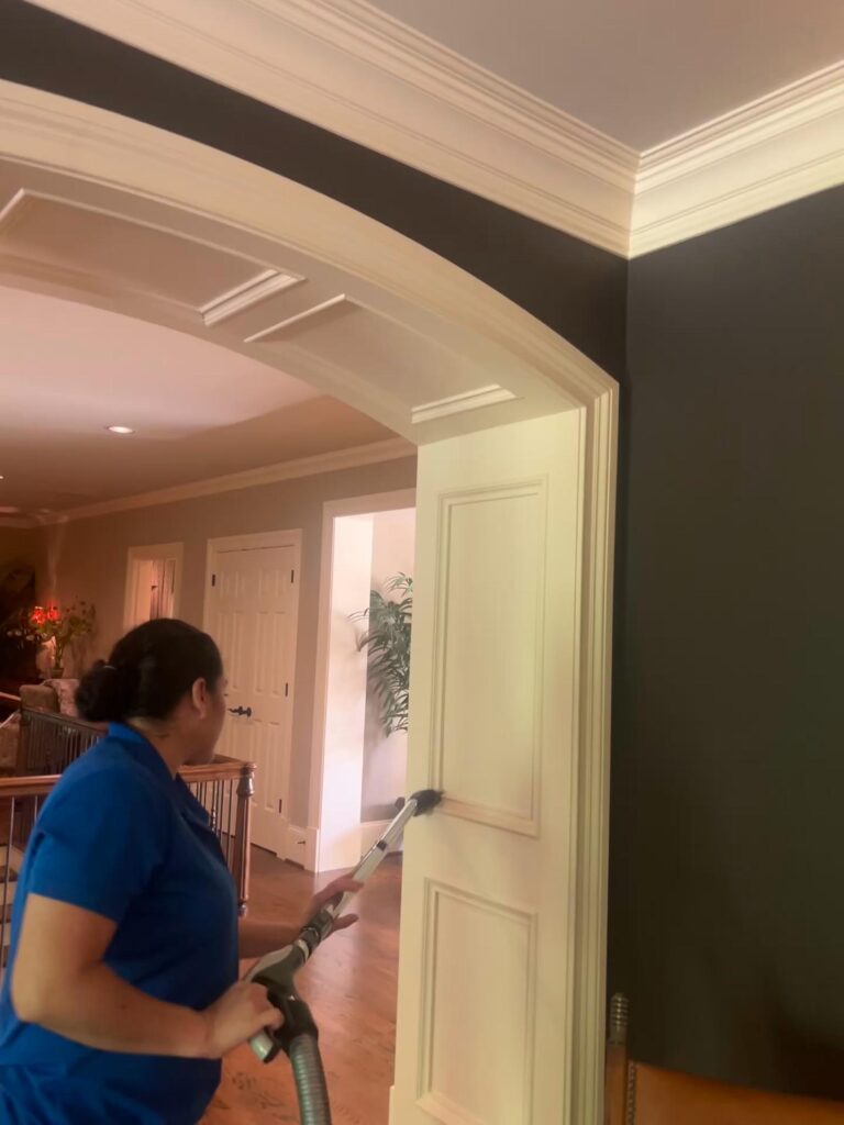 A woman in a blue shirt uses a vacuum cleaner with a brush attachment to clean a white door in a house.