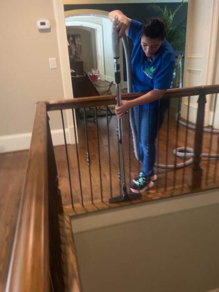 A person in a blue shirt is vacuuming the wooden floor of an upstairs hallway near a railing, showcasing professional cleaning services in action.