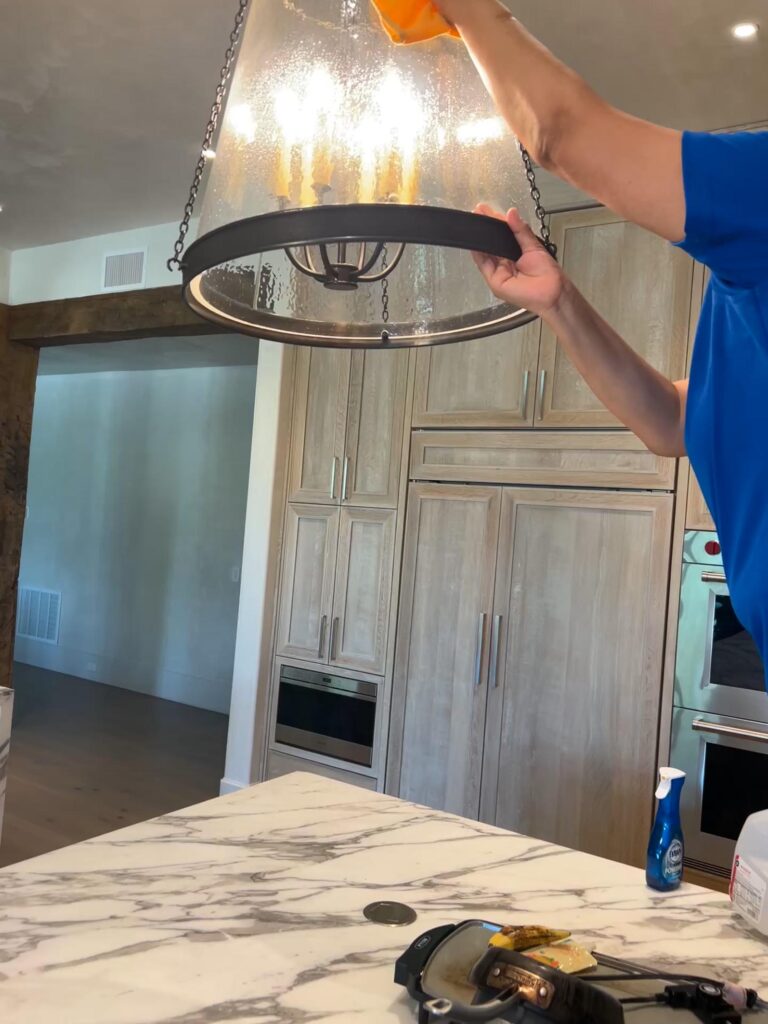 Person in a blue shirt cleaning a glass pendant light fixture above a marble kitchen counter, with cleaning supplies visible nearby.
