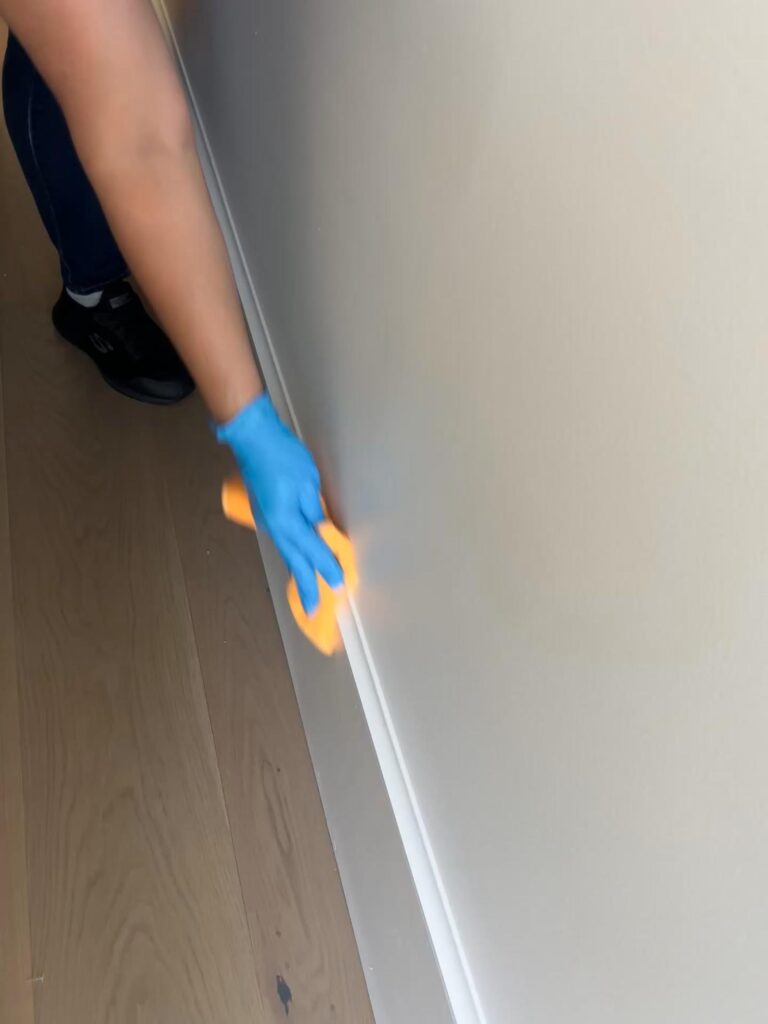 A person wearing a blue glove uses an orange cleaning tool to wipe the baseboard along a light-colored wall and wooden floor, demonstrating the attention to detail provided by professional cleaning services.