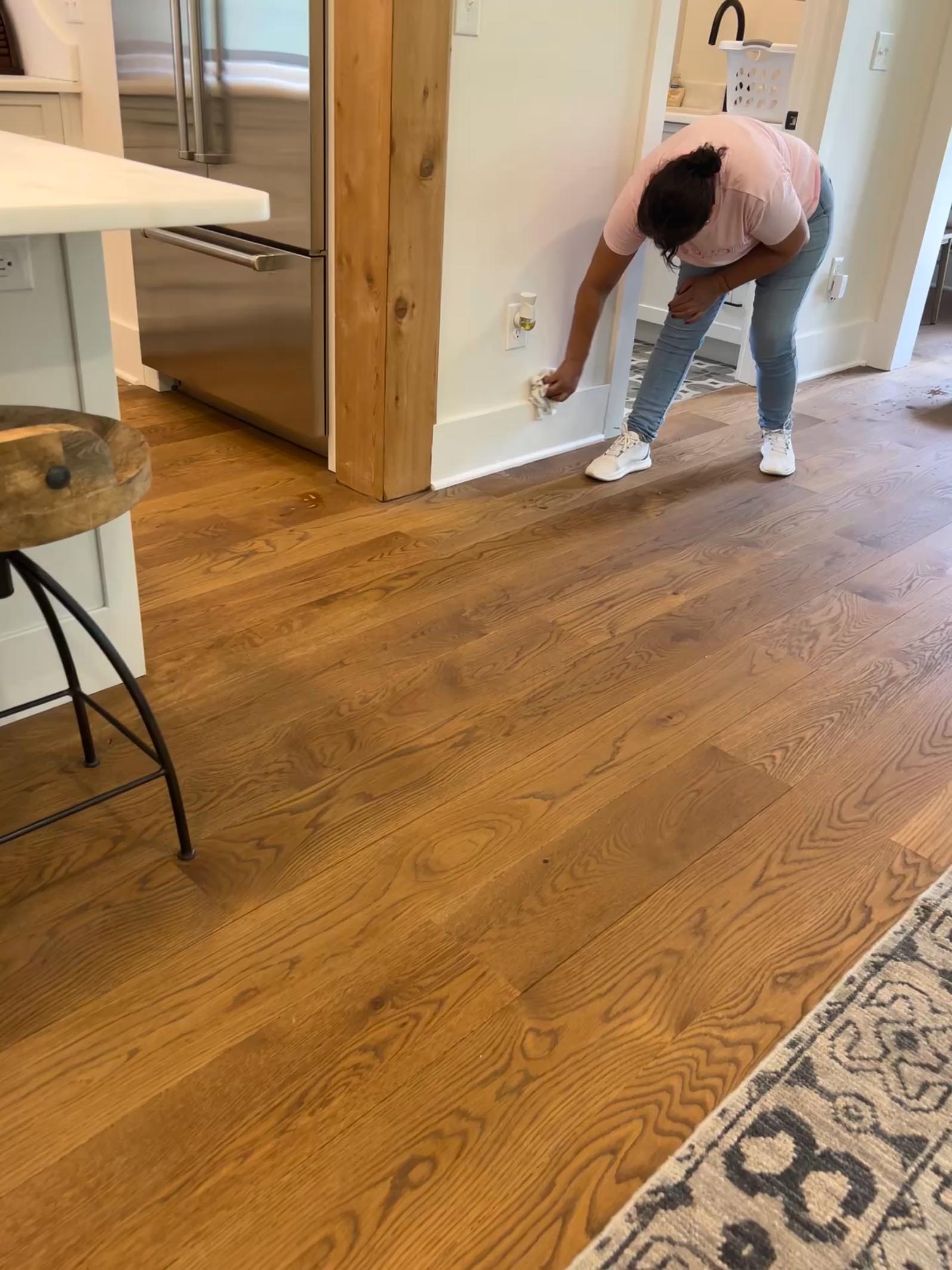 A person in a pink shirt and jeans bends down to plug something into a wall outlet in a kitchen with wooden floors.