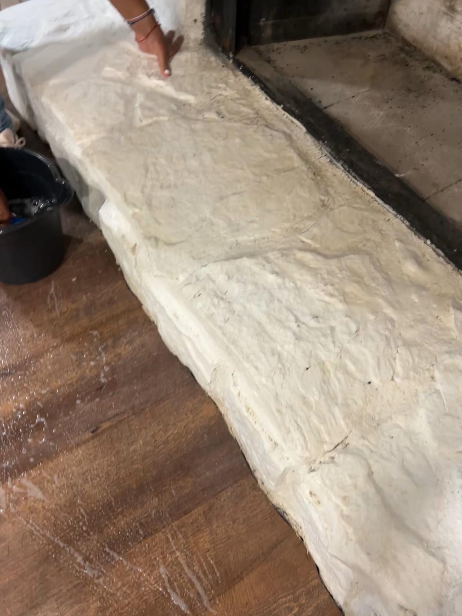 A person spreads white plaster onto a stone or concrete ledge beside a fireplace, with cleaning services tools and a bucket nearby on the wooden floor.