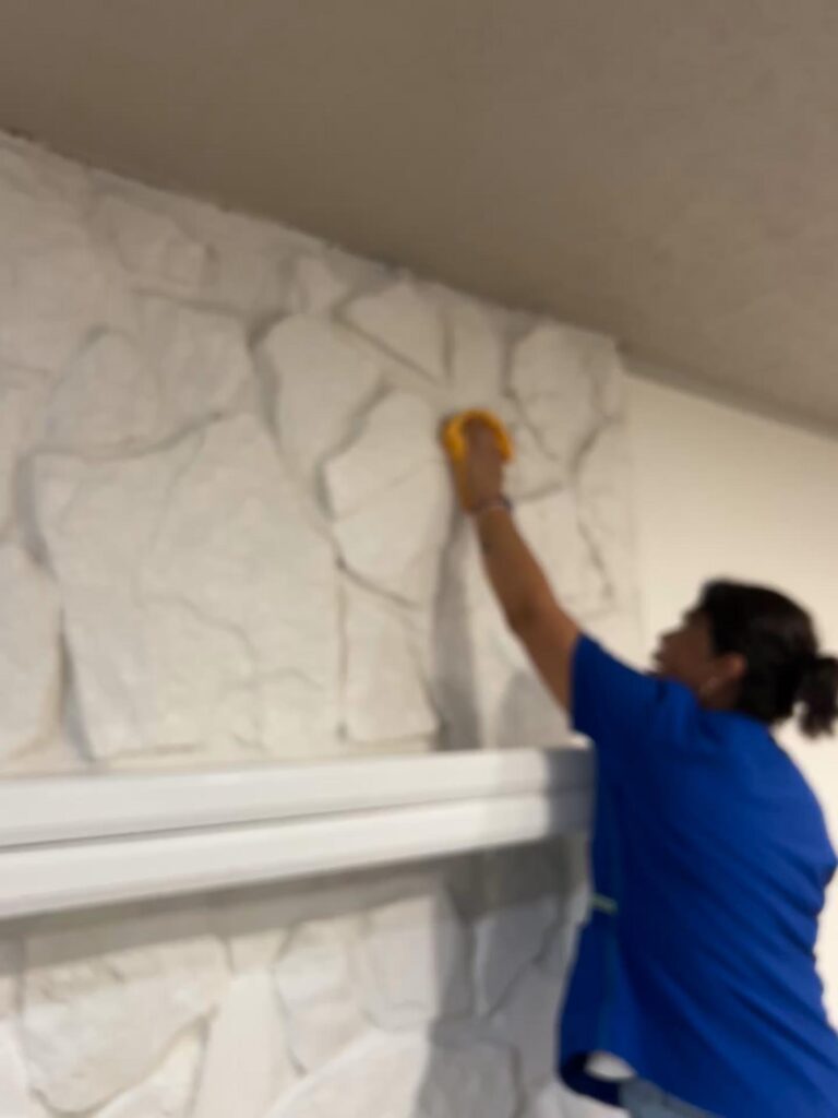 A person in a blue shirt reaches up to clean a white stone wall with a yellow cloth, showcasing professional cleaning services.