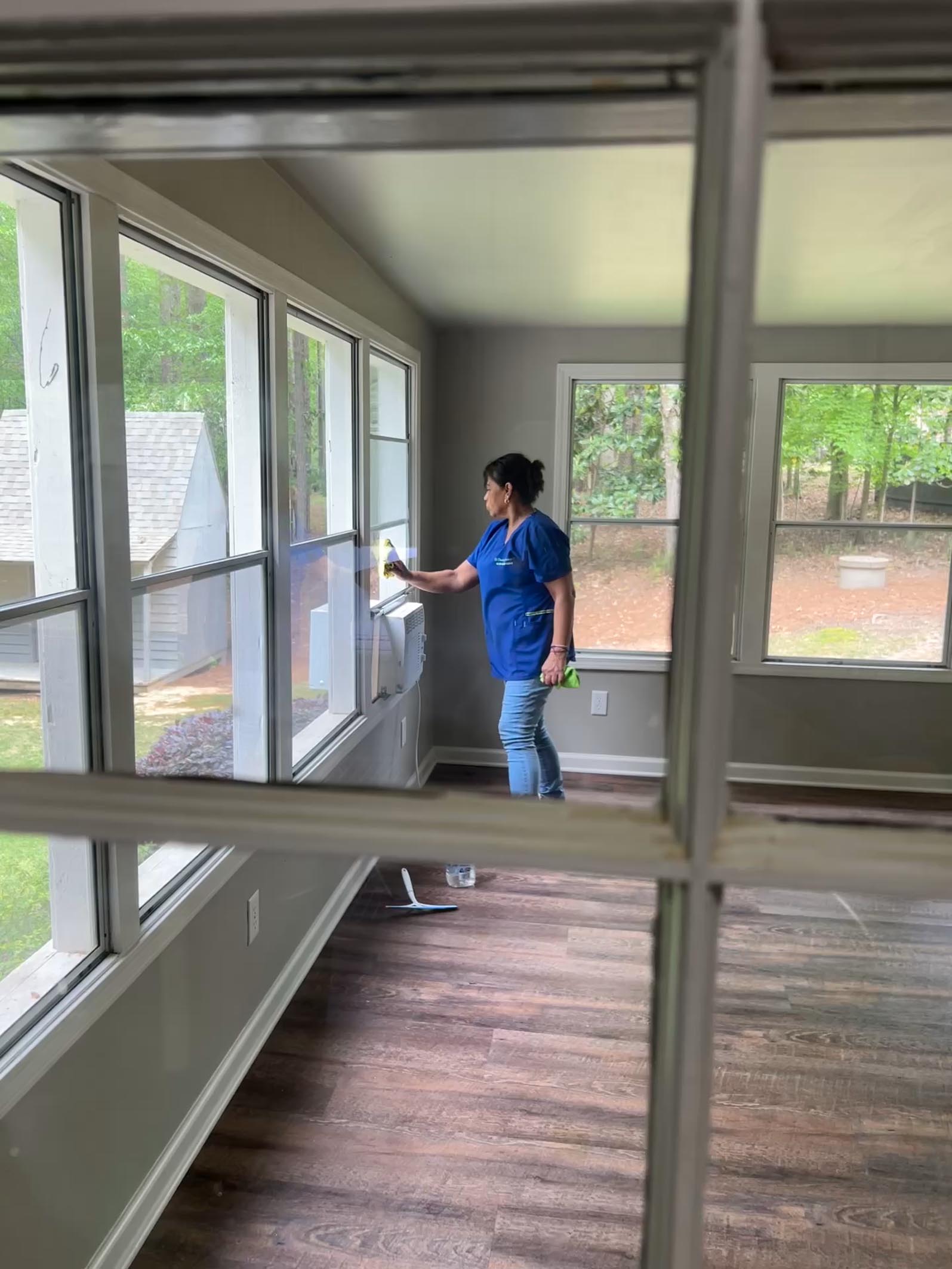 A woman in a blue shirt and jeans cleans the inside of a window in a bright, empty room with large windows and wood floors, suggesting demolition cleaning in Georgia.