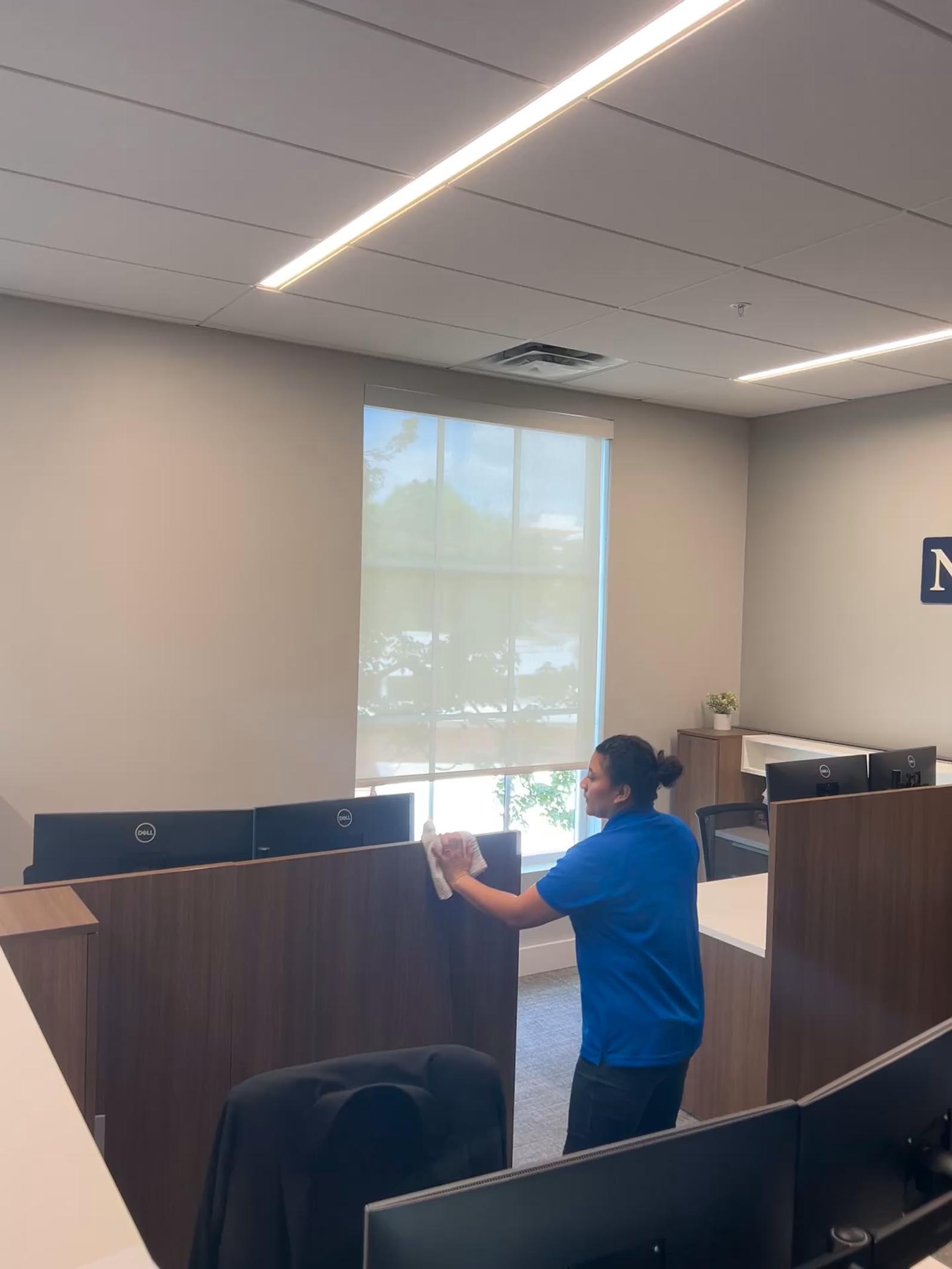 A person in a blue shirt from cleaning services tidies an office desk in a modern workspace with several computer monitors and a large window.