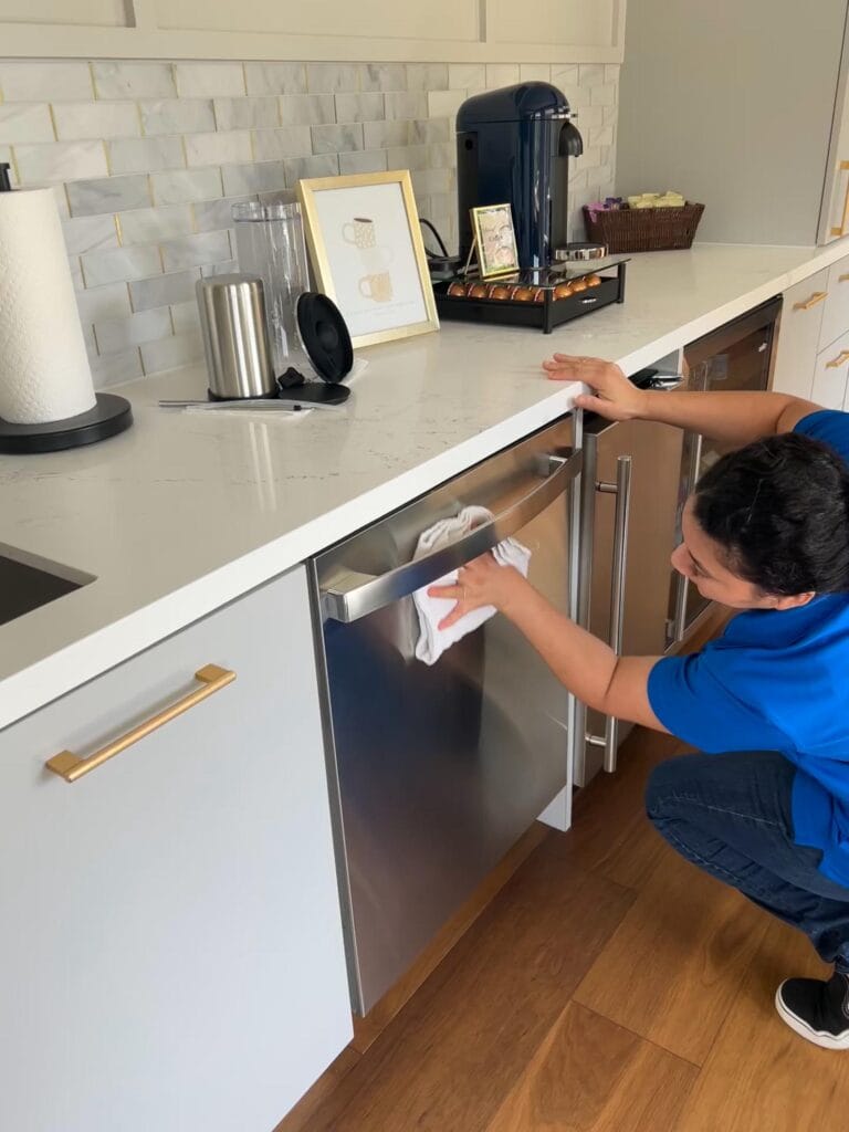 A person in a blue shirt is cleaning a stainless steel dishwasher in a modern kitchen with white cabinets and a coffee station on the counter. This image is about CK cleaning.