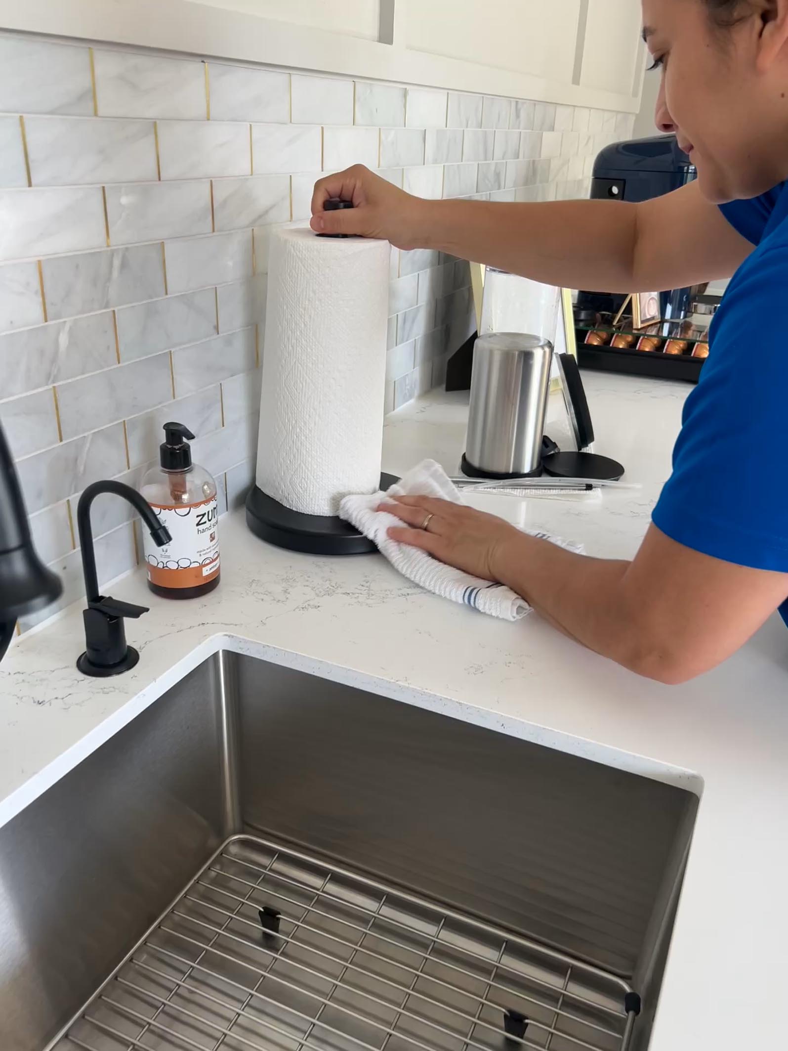 Residential cleaning in Marietta: A person in a blue shirt cleaning a white kitchen counter with a towel, near a paper towel holder, soap dispenser, and sink.