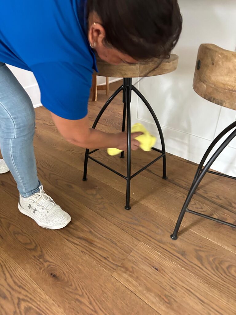 A person in a blue shirt wipes the metal legs of a wooden stool with a yellow cloth on a wooden floor.