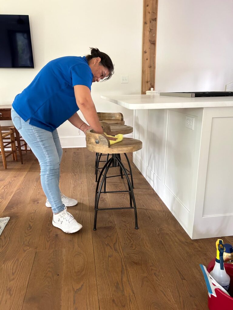 One-Time Cleaning in Georgia: A woman wearing a blue shirt and jeans cleans a wooden barstool with a yellow cloth in a modern kitchen with white cabinets and wood flooring.
