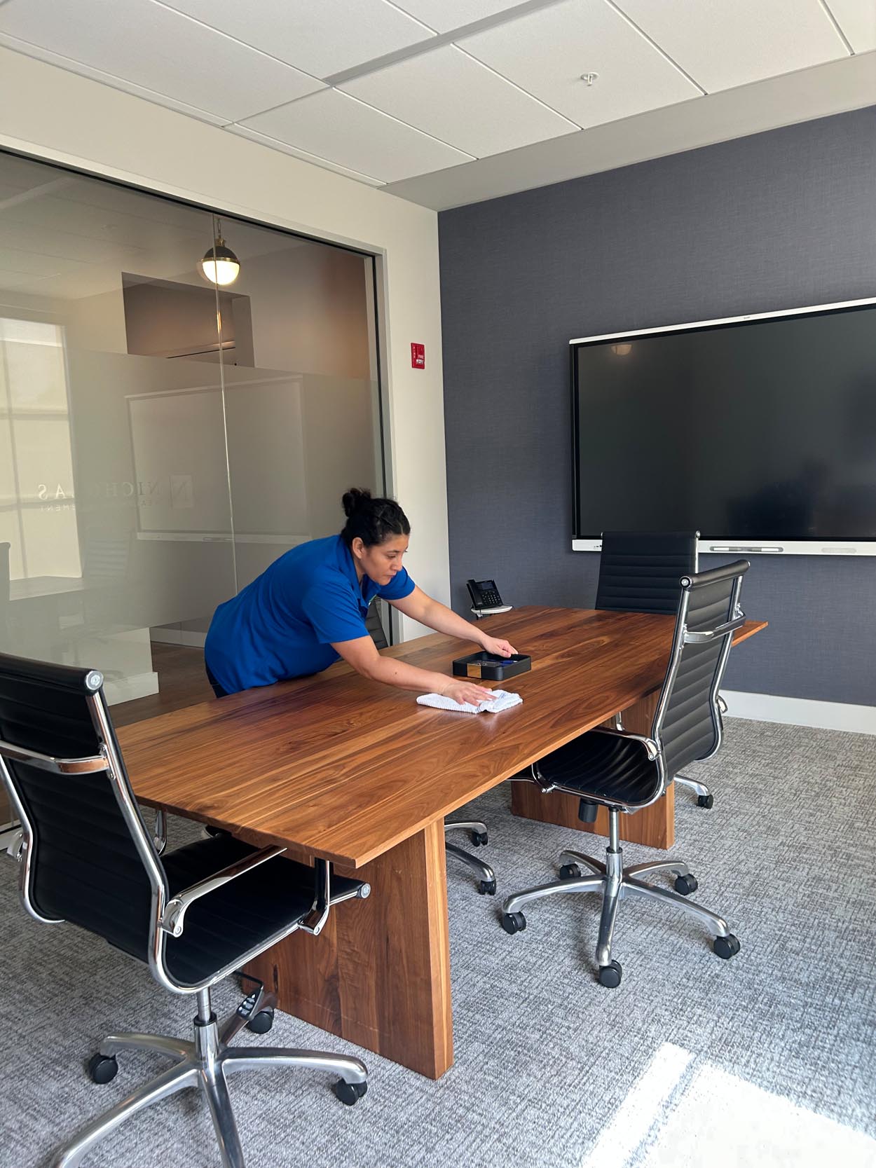 Commercial Cleaning in Georgia: A CK Cleaning staff in a blue shirt is cleaning a conference table in a modern office meeting room with four chairs and a large wall-mounted screen.