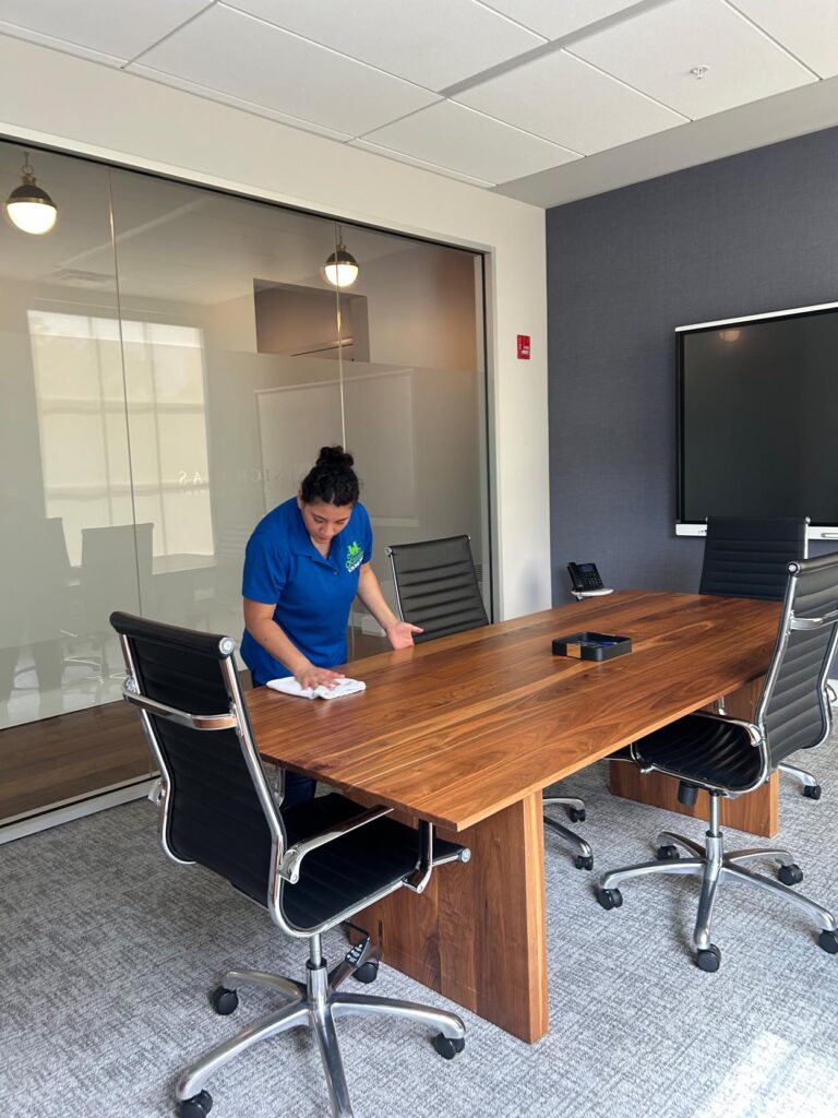A person in a blue shirt is cleaning a wooden conference table in a modern office meeting room with black chairs and a large wall-mounted screen.