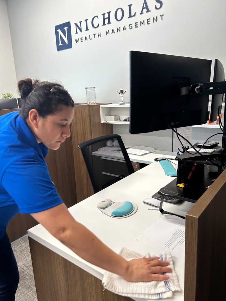 A woman in a blue shirt wipes down a desk in an office with a "Nicholas Wealth Management" sign on the wall.