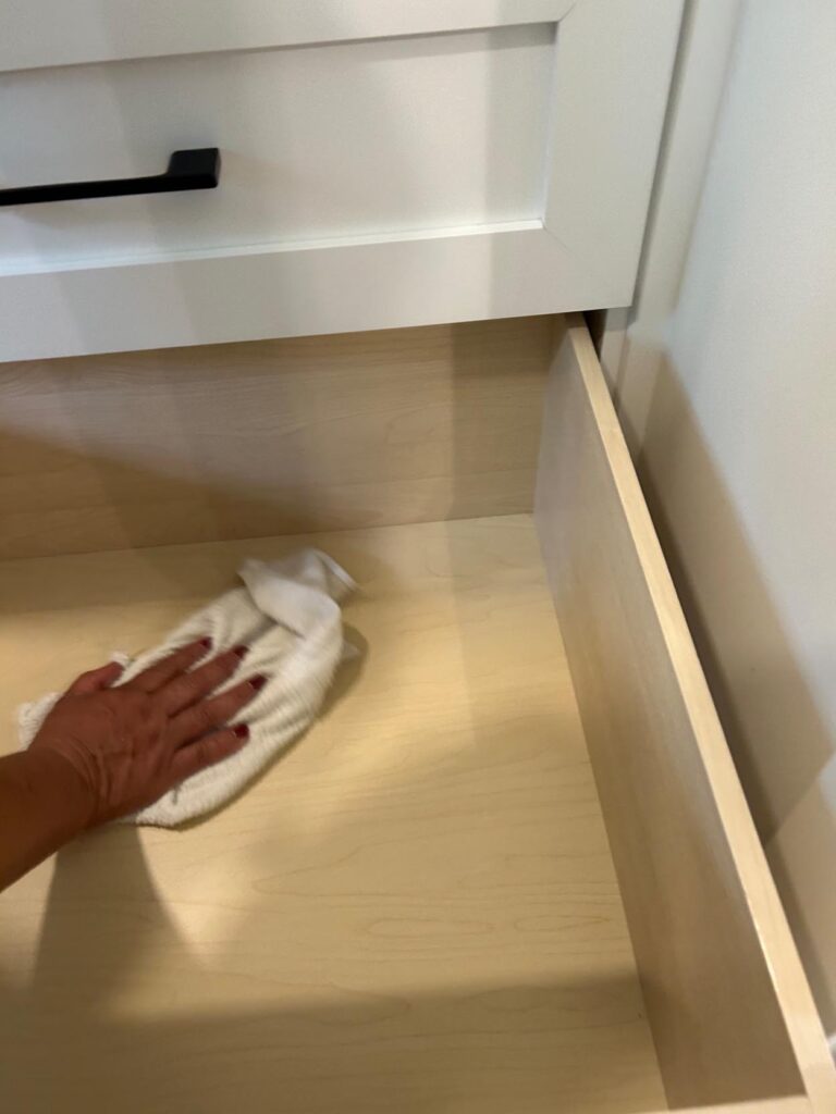 A hand is wiping the inside of an empty wooden drawer with a white cloth.