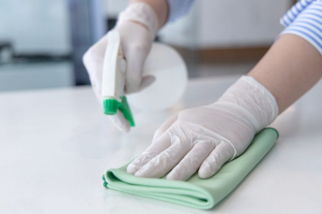 A person wearing gloves sprays a cleaning solution onto a surface and wipes it with a green cloth during post-construction cleaning.