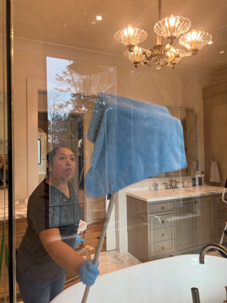 A woman wearing gloves uses a large blue cloth on a pole to clean a glass shower wall in a modern bathroom.