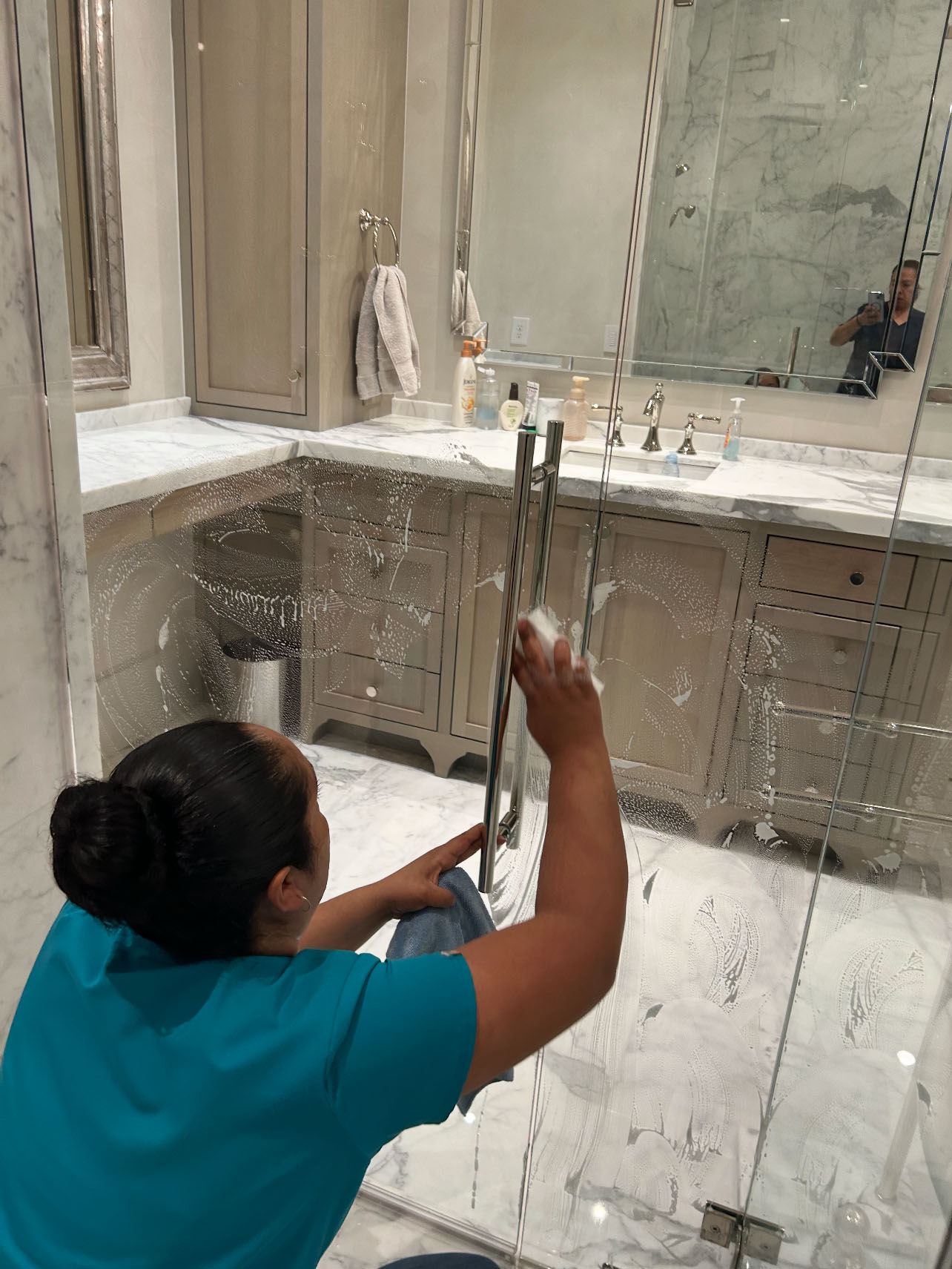 Careers with CK Cleaning: A person in a teal shirt cleans a glass shower door with soap in a marble bathroom; another person is visible in the mirror taking the photo.