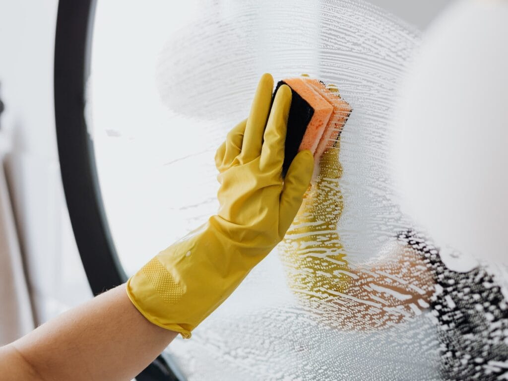 A person wearing yellow rubber gloves cleans a glass surface with a sponge and soapy water, perfect for thorough move-in move-out cleaning services.