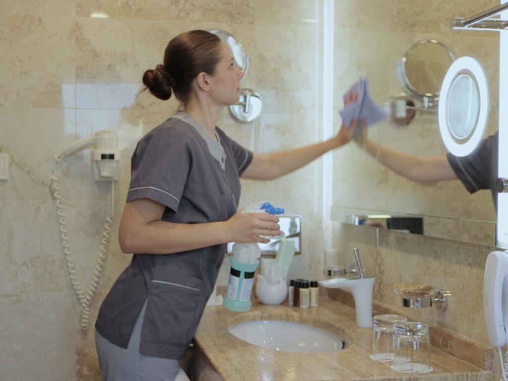 A person in cleaning uniform wipes a bathroom mirror with a cloth while holding a spray bottle, showcasing move-in move-out cleaning services by the sink on a marble countertop.