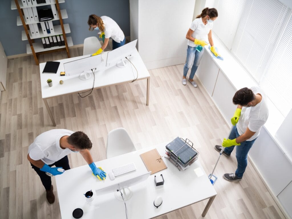 Four people in white shirts and yellow gloves provide move-in move-out cleaning services, wiping desks, mopping floors, and cleaning windows in a bright office workspace.