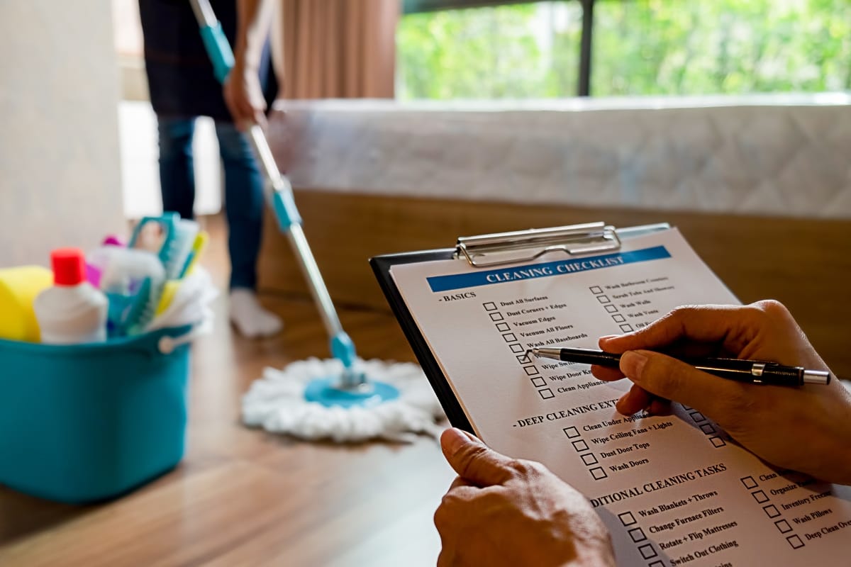 A person marks a weekly cleaning checklist on a clipboard while another mops the floor near a bed; a blue bucket with cleaning supplies is in the foreground.