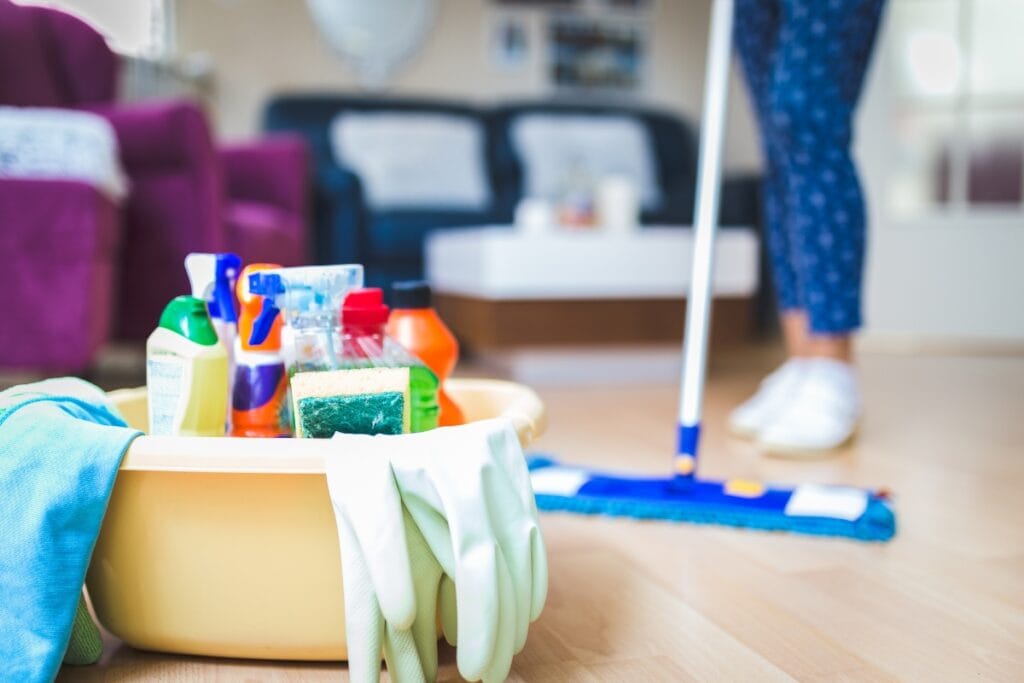 A plastic tub with cleaning supplies and gloves sits on the floor; a person in the background is mopping a living room, following their weekly cleaning checklist.