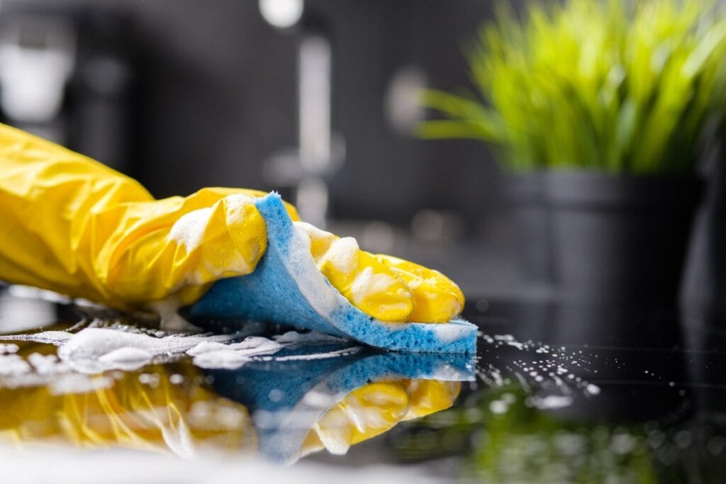A hand in a yellow glove wipes a soapy surface with a blue sponge, following a weekly cleaning checklist; green plants are visible in the background.