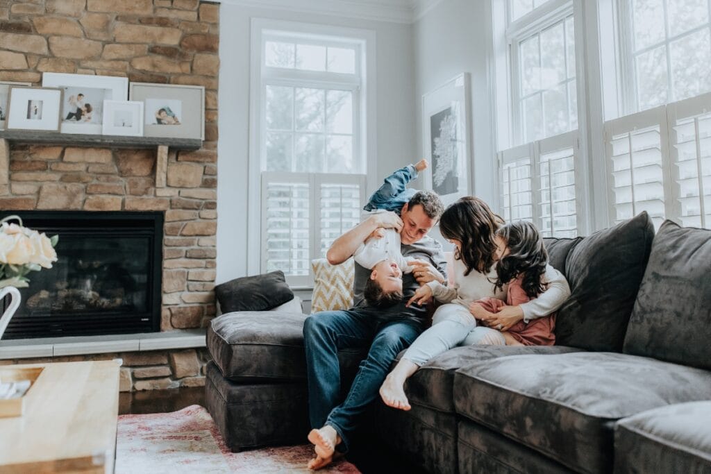 A family of four sits on a gray sectional sofa in a living room, playfully interacting and laughing together near a fireplace and large windows, enjoying their spotless space thanks to a weekly cleaning checklist.