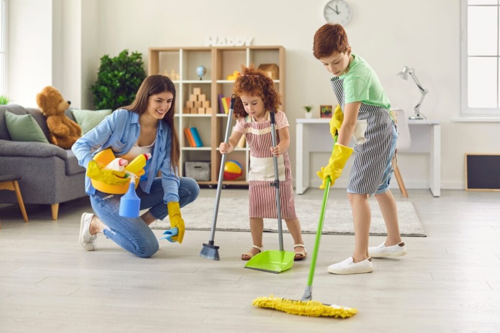 A woman and two children follow their weekly cleaning checklist as they tidy the living room; one sweeps, one mops, and the woman holds cleaning supplies. All are wearing rubber gloves.