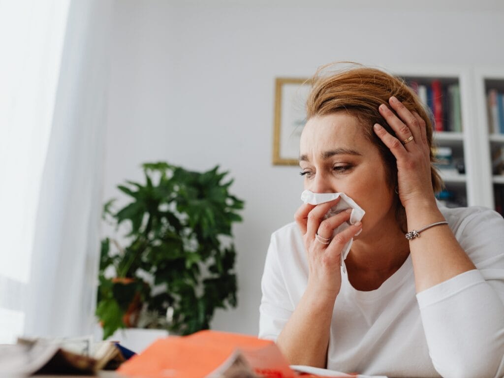 A woman holding a tissue to her nose sits at a table, looking unwell and concerned—an all-too-familiar scene for allergy sufferers—with papers and a plant visible in the background.