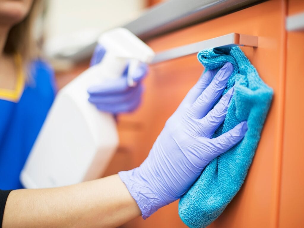 A person wearing purple gloves cleans an orange cabinet handle with a blue cloth and holds a spray bottle—an essential step in residential cleaning, especially for allergy sufferers.