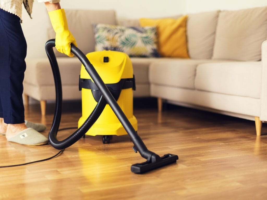 Person wearing yellow gloves uses a yellow vacuum cleaner to clean a wooden floor in a living room, showcasing the quality of Residential Cleaning Services Near Me, with a beige sofa and decorative pillows in the background.