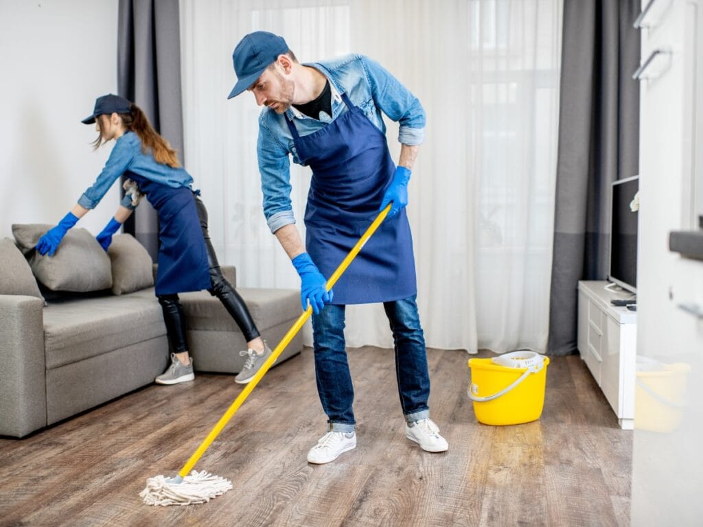 Two cleaners in blue aprons and gloves clean a living room; one mops the wooden floor while the other wipes down a sofa. A yellow bucket is on the floor, showcasing the quality you can expect from Residential Cleaning Services Near Me.