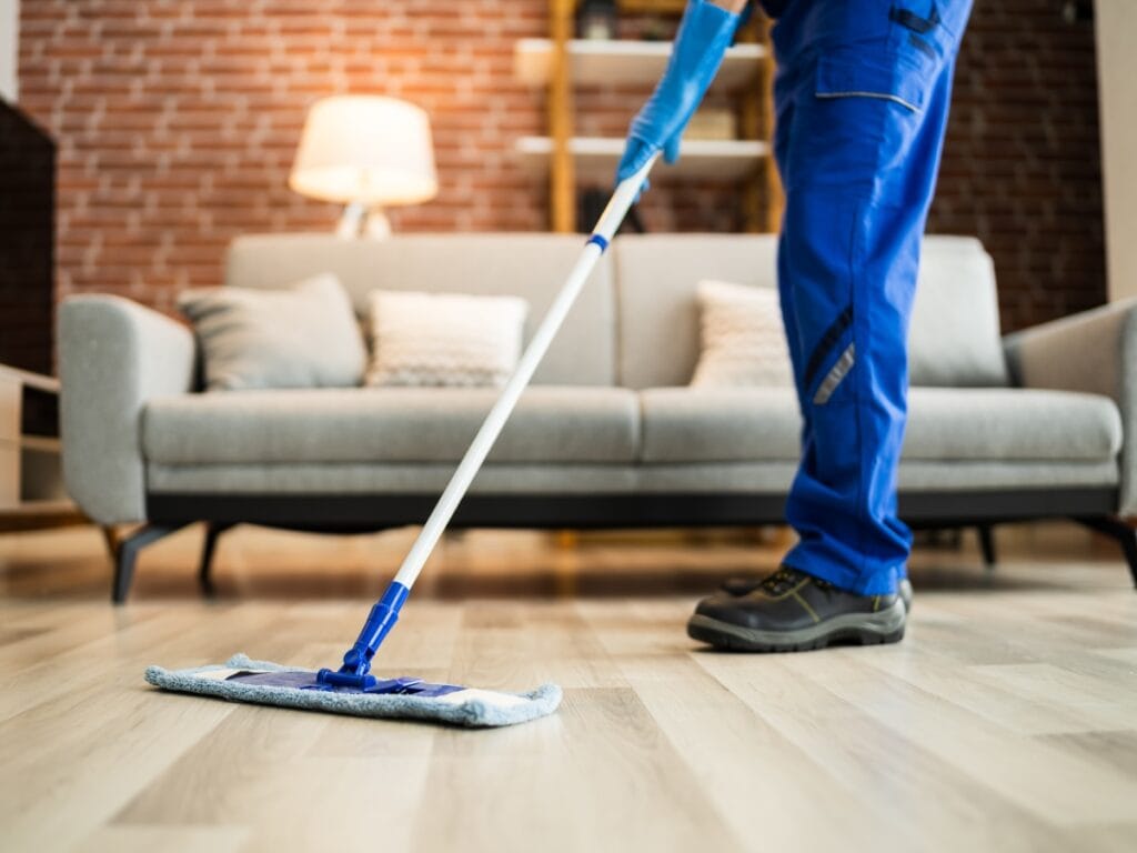 A person wearing blue overalls and gloves is mopping a wooden floor in a living room, showcasing the quality of Residential Cleaning Services Near Me, with a cozy sofa and lamp in the background.
