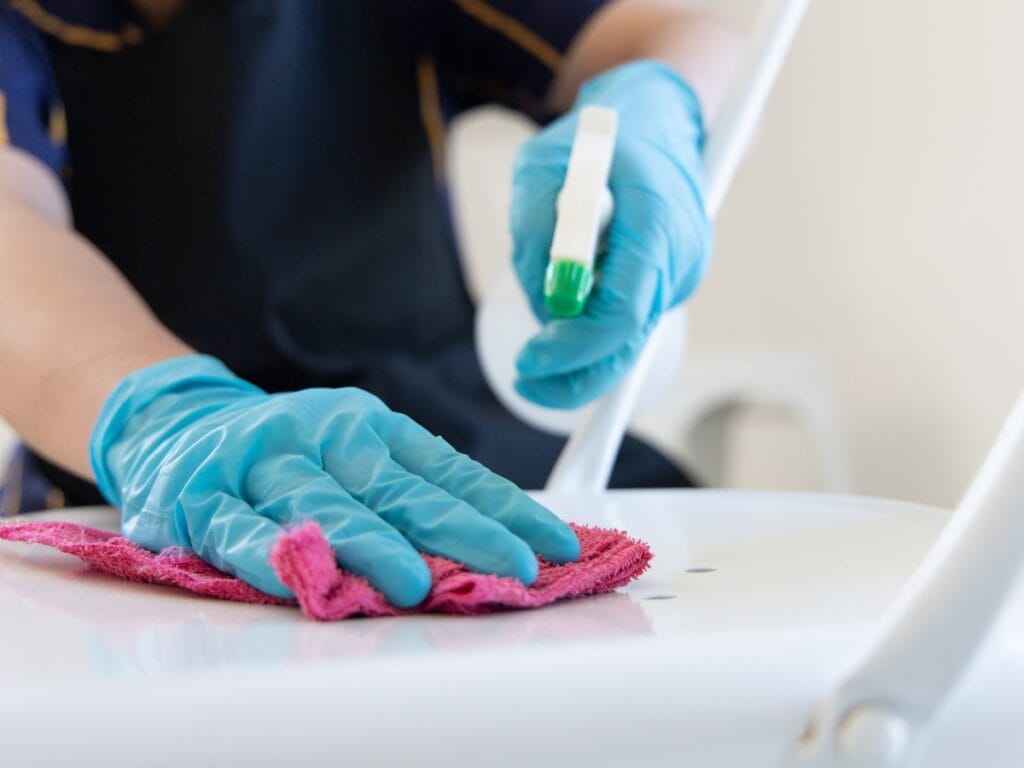 Wearing blue gloves, a person uses a spray bottle and pink cloth to clean a white chair, demonstrating the detailed care you might expect when learning how do cleaning services charge for thorough cleaning.