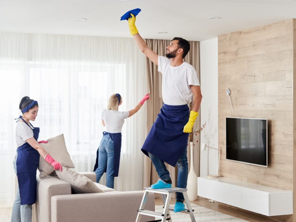 Three people in aprons and gloves clean a modern living room; one wipes a ceiling fixture while standing on a ladder, another cleans a window, and the third arranges cushions—demonstrating the thorough approach often considered in How Do Cleaning Services Charge.