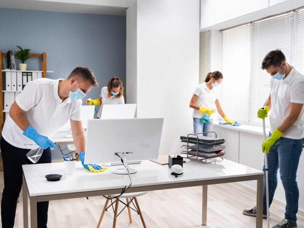 Four people wearing masks and gloves clean an office, wiping surfaces and mopping the floor around desks and computers, showcasing the thorough work you can expect when learning how do cleaning services charge.