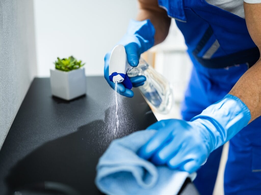 Person wearing blue gloves and overalls cleaning a surface with a spray bottle and cloth; if you’re unhappy with cleaning services, trust a professional who cares. Small potted plant in the background.