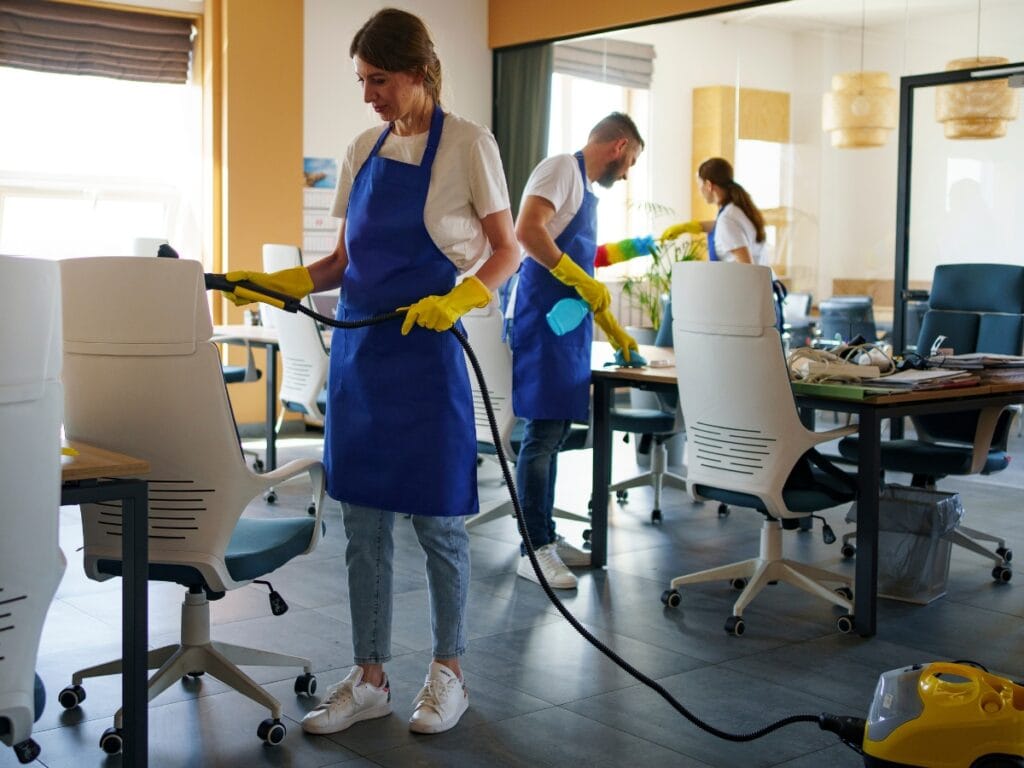 Three people wearing blue aprons and yellow gloves are cleaning an office space, wiping desks and vacuuming the floor—perfect for those who are unhappy with cleaning services that don't meet high standards.