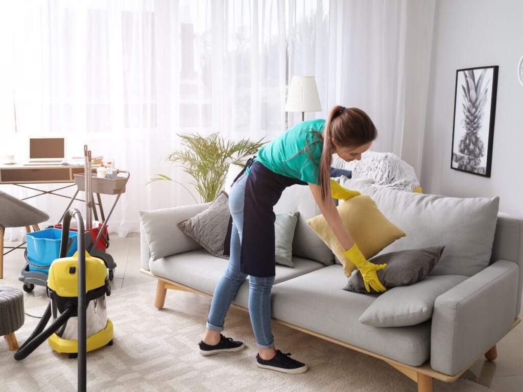 A woman wearing gloves and an apron arranges pillows on a sofa in a tidy living room with cleaning supplies nearby, showcasing the results of deep cleaning vs regular cleaning.