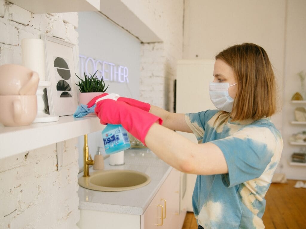 A person wearing a mask and pink gloves deep cleans a kitchen shelf with a spray bottle and cloth, highlighting the difference between deep cleaning vs regular cleaning.