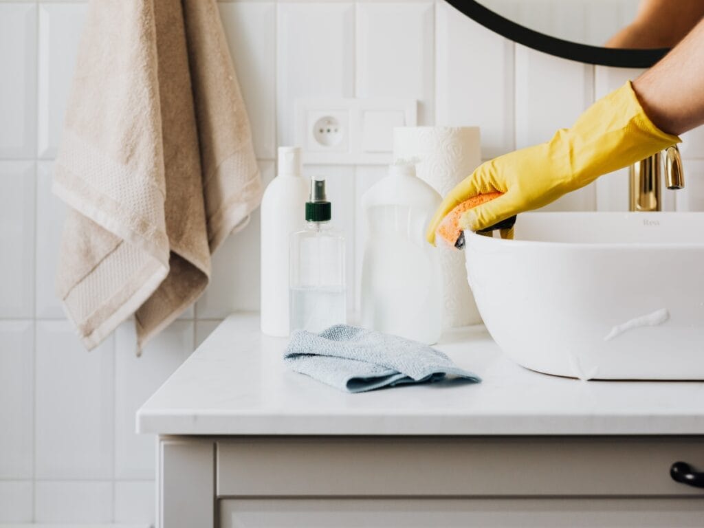 A person wearing a yellow glove cleans a white bathroom sink with a sponge, highlighting the difference between deep cleaning vs regular cleaning; cleaning supplies and towels are visible on the countertop.