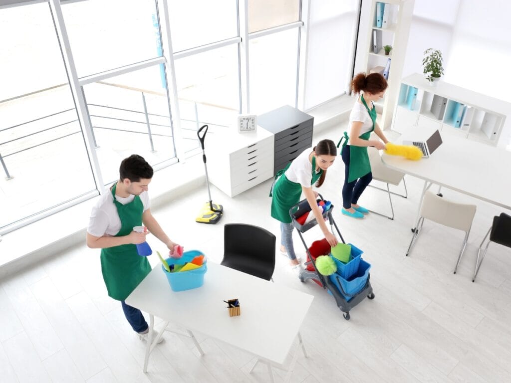 Three people in green aprons clean an office with supplies; one wipes a table, another uses a laptop&mdash;perhaps to schedule house cleaning&mdash;and the third pushes a cart, showcasing professional house cleaning in action.