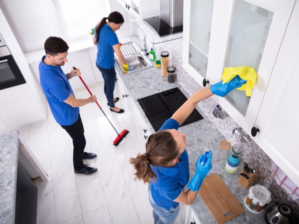 Three people in blue shirts deliver professional house cleaning in a modern kitchen; one sweeps the floor, another wipes a cabinet, and the third washes dishes at the sink.